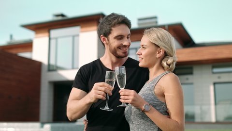 Caucasian man with blonde woman hugging outdoor. Portrait of romantic couple drinking champagne near luxury apartment. Rich people toasting champagne outside villa.