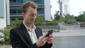 Side view of happy young Caucasian man texting on phone outside business office, smiling. Lifestyle, communication concept
