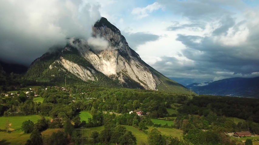 Aerial, tracking, drone shot, over buildings and green fields, overlooking a bare, steep mountain, in the Swiss alps, at golden hour, on a partly sunny day, in Leuk, Vallis, Switzerland