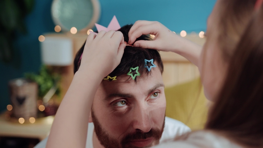 Portrait of miserable upset daddy crying for help while playing with his daughter. Poor tired father with hairpins in hair dressed like princess sitting with his child on bed.