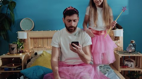 Portrait of exhausted young parent father using a smartphone sitting on bed and touching his face. Handsome young man is tired of playing with kids. Fancy cute girl dancing and having fun on bed
