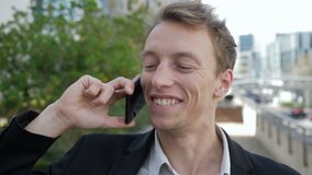 Front view of happy young Caucasian man talking on phone outside business office, smiling. Lifestyle, communication concept
