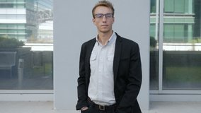 Front view of serious young Caucasian man in spectacles standing outside business office, thinking, correcting hair, buttoning jacket, crossing hands. Lifestyle concept