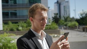 Side view of happy young Caucasian man texting on phone outside business office, laughing, looking satisfied. Lifestyle, communication concept