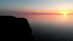 Adventurous man on top of the mountain on the sea shore enjoying the beautiful view during a vibrant sunset. Beautiful natural landscape at sunset aerial video