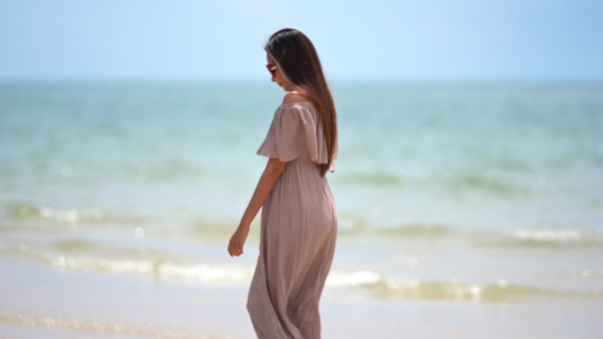 A beautiful Asian woman wearing large sunglasses dressed in a pale pink flowing dress walks along the beach while the waves lap against? her bare feet.