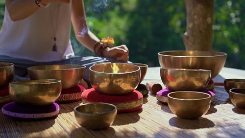 Superslowmotion shot of a woman master of Asian sacred medicine performs Tibetan bowls healing ritual. Meditation with Tibetan singing bowls. She sits in a gazebo for meditation with a beautiful