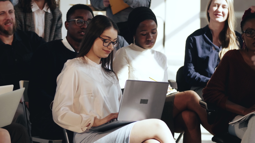 Happy smiling young Caucasian business woman sitting in multiethnic audience, clapping to speaker at conference seminar.