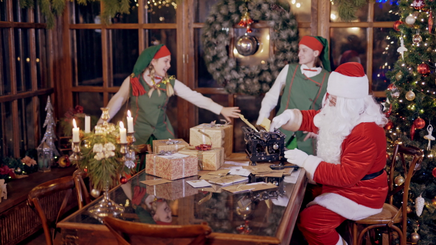 Merry atmosphere with elves and Santa Claus. Santa with white beard in red costume is typing letter on the background of funny elves. New Year