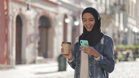 Young muslim woman wearing hijab headscarf walking in the city center, using smartphone and drinking coffee. Communication, online shopping, social network concept.