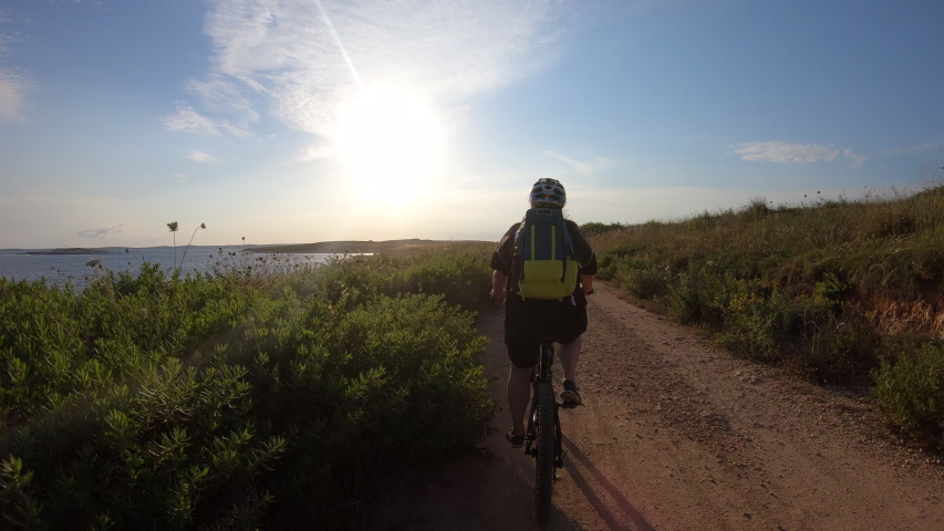 Right behind tracking shot of a female mountain biker riding along the coastline on a dusty trail in sunshine