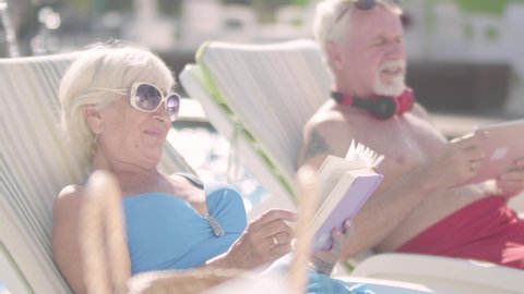 Cute couple lying on sunbeds near the pool. Mature woman reading a book while old man looking at a tablet. Happy loving family. Rest in hotel.
