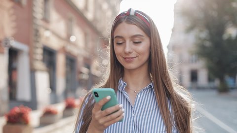 Smiling young woman wearing in blue and white striped dress shirt walking around old street using smartphone. Communication, social networks, online shopping concept.