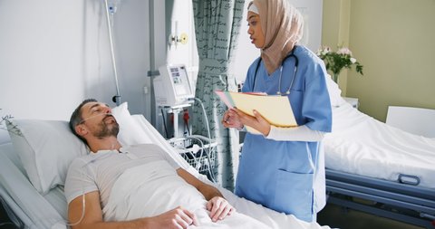 Young mixed race female doctor wearing hijab holding files and talking to a middle aged Caucasian male patient lying in hospital bed. Healthcare workers in the Coronavirus Covid19 pandemic