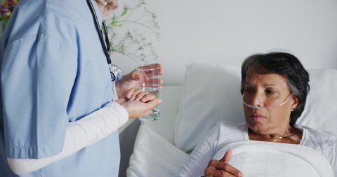 Close up of senior mixed race woman lying in hospital bed taking pills with water, assisted by a female healthcare worker seen in mid section. Healthcare workers in the Coronavirus Covid19 pandemic
