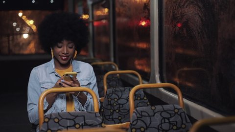Attractive young african american woman using smartphone riding at public transport and looking out the window. Night time. City lights background.