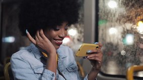 Smiling young african american woman watching funny video on the smartphone riding in the public transport. Night time. Close-up. City lights background.