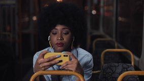 Young african american woman watching video on the smartphone riding in the public transport. Night time. Close-up. City lights background.