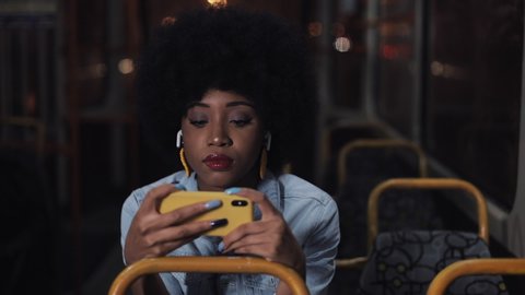 Young african american woman watching video on the smartphone riding in the public transport. Night time. Close-up. City lights background.