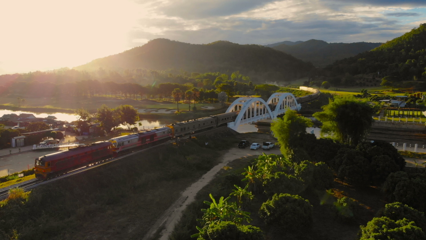 Aerial view of Unseen locomotive passenger train run through the old white bridge during sunset or sunrise with flare light. Tha Chom Phu white bridge a tourist attraction famous of Northern Thailand Royalty-Free Stock Footage #1033273538