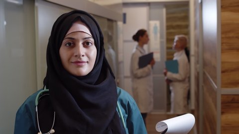 Close up of young Muslim female medical worker standing with outpatient card, looking at camera while her colleagues talking on background