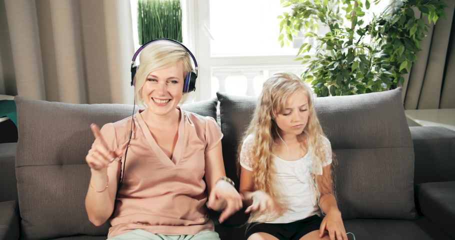 Middle-aged woman and teenage girl having fun together, listening to favorite soman and making dancing movements while sitting on sofa
