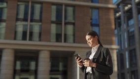 Waist up panning shot of smiling young woman in formal suit standing outside with takeaway coffee cup and text messaging on cell phone. Businesswoman looking away dreamily while thinking of response - Powered by Shutterstock - Get 15% off with code: PIKWIZARD15