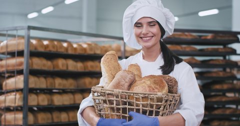 Smiling pretty lady baker holding a basket with fresh baked bread in a bakery factory