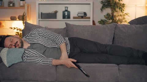Portrait of person handsome guy napping on sofa at home at night with remote control in hand, TV clicker is falling on floor. People and lifestyle concept.