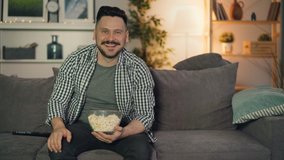 Cheerful person watching funny show on TV laughing pointing at screen eating popcorn sitting on couch in modern apartment. People and fun concept.