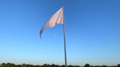 White flag waiving in the wind. Moon is visible in the background against a dark blue sky with no clouds. Small number of trees is visible on the horizon. Symbol of surrender, flag pole, sun kissed