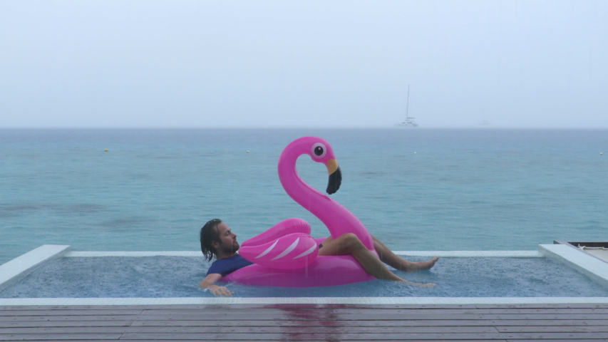 Funny fail video in vacation rain of holidays getaway travel raining away under heavy rainfall with man falling while trying to get out of flamingo float in luxury pool with funny face. Royalty-Free Stock Footage #1033325633