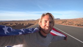 Man on road takes selfie while holding American flag; road trip concept; Young man standing on empty highway road in USA taking selfie video