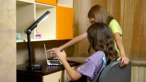 Two girls girlfriends, teenagers students sit together at the training table and use a laptop in the evening. they are cheerful and happy