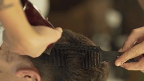 The barber trims skillfully client's hair from the back using haircut machine and a comb. It is a close up video of vertical orientation. The background is blurred.