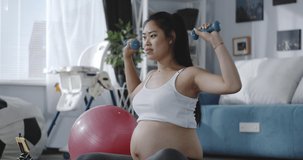 Medium shot of a young pregnant woman watching a tutorial video while training with dumbbells