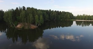 4K aerial summer early morning high quality video footage of pristine lush green pine tree surrounded forest lake with dark calm water reflecting clouds, a dam, island and a sand beach near small town