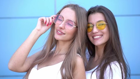 Two young beautiful smiling hipster girls in trendy summer colorful neon sunglasses. Women posing near blue wall