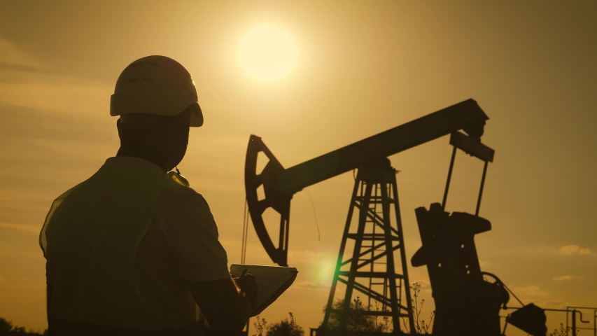 Silhouette of man engineer with clipboard and securities for the oil pumping unit overseeing the site of crude oil production at sunset.