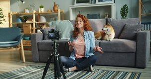 Slow motion of female vlogger attractive young lady recording video at home with cute dog sitting on floor in apartment talking and gesturing. People and pets concept.