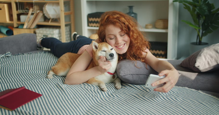 Happy teenage girl is taking selfie with adorable dog kissing pet using smartphone camera at home lying on couch together. People and affection concept.