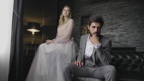 slow motion bearded man with chin propped in hand and lady in long dress sit on leather sofa and chair like old photo