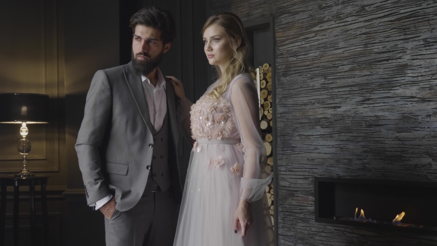 Slow motion elegant blond lady in nice dress and bearded man in grey suit stand at fireplace in dim room | Shutterstock HD Video #1033402967