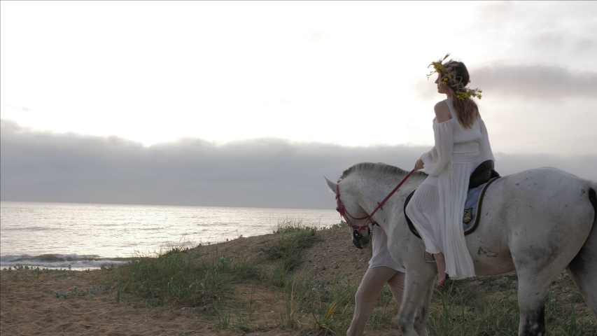 brunette in long white summer dress rides horse and man holds halter walking along sand beach at sunset slow motion