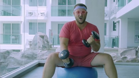 Playful fat man in a pink T-shirt and pink glasses is engaged in fitness with dumbbells and a fit ball in the gym. 