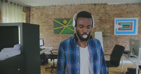 Young black man blows bubbles with a chewing gum, listens to music in headphones and leaves office while his colleagues discussing graphs on screen on blurred background Stock Video