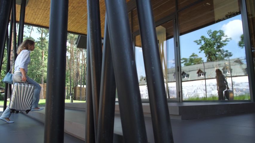 Back view of woman carrying suitcase and entering hotel, buildings of hotel reflected in wide glazed entrance to hall. Pretty casually dressed female spending weekend leisure in countryside