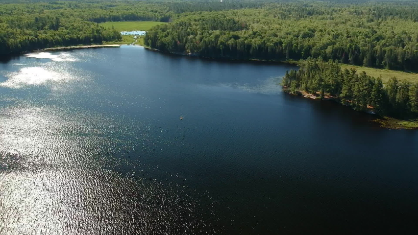 Aerial shot flying forward high above lake and gradually descending to the water with sun glares and kayak canoe boat crossing the lake from right to left. Grundy lake, Northern Ontario, Canada.