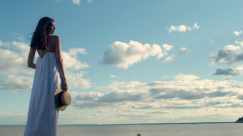 beautiful slender girl in long white dress holds straw hat in her hand, shakes long hair, turns  face into camera. woman smiles and straightens her hair. The view from the back. Warm sunset on the sea