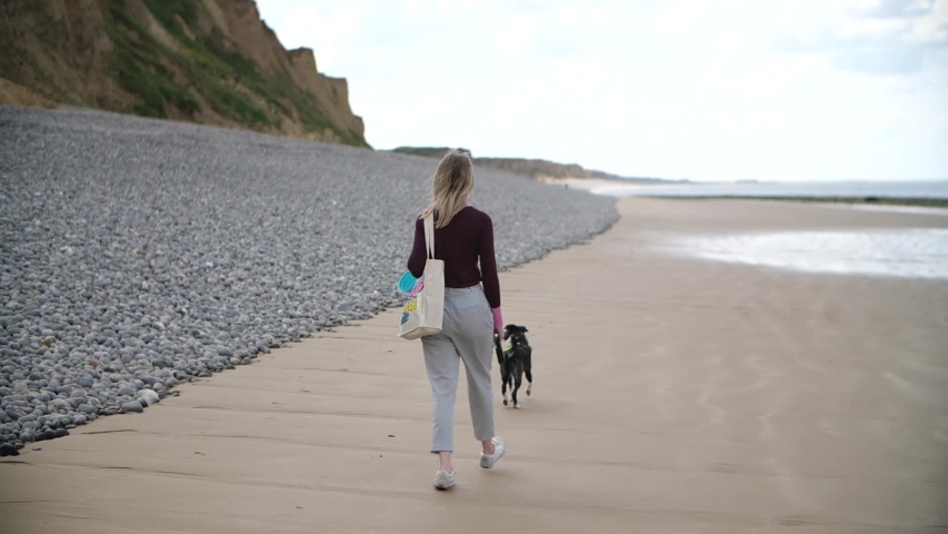 Slow mo shot of girl walking her dog along a sandy beach. The woman has light blonde hair that blows in the wind, grey trousers and is wearing a nice purple jumper with a tote bag 
